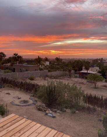 Un magnifique coucher de soleil peint le ciel en orange et rose vibrants sur un paysage désertique. Des palmiers, des buissons, des cactus et des maisons sont disséminés dans le paysage.