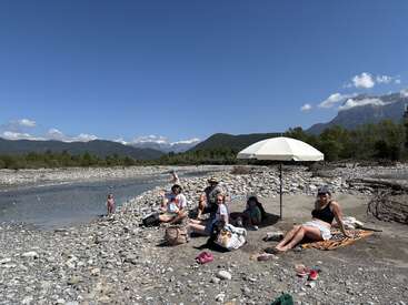 Un grupo de personas se relajan bajo una sombrilla blanca en la orilla rocosa de un río, rodeados de montañas, cielo despejado y enseres dispersos, disfrutando de un día soleado al aire libre.