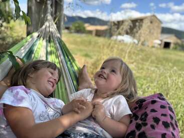 Dos niños pequeños se relajan juntos en una hamaca verde al aire libre, compartiendo risas y cogidos de la mano. Detrás de ellos hay un campo soleado, edificios rústicos y montañas.