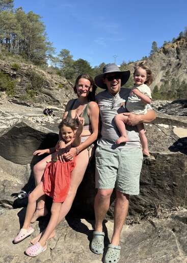 A smiling family of four enjoys sunny weather outdoors, sitting together on large rocks, surrounded by trees and mountains, radiating happiness and togetherness during their outing.