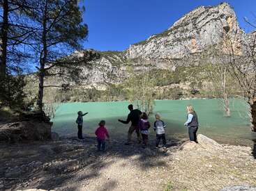 A group of adults and children stand by a turquoise lake, surrounded by trees and dramatic rocky mountains under a clear, bright blue sky. Nature outing.