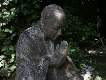 A imagem apresenta uma estátua de pedra desgastada de um homem careca em uma pose de oração, tendo como pano de fundo uma vegetação exuberante e algumas pedras brancas.