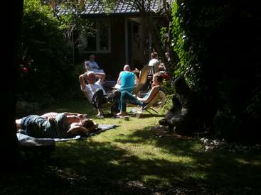 A imagem mostra um grupo de pessoas relaxando em um quintal, com algumas sentadas em cadeiras e outras deitadas na grama, cercadas por árvores e uma casa ao fundo.