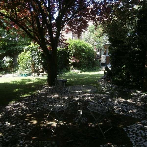 A imagem mostra uma cena serena de jardim, com uma mesa e cadeiras em primeiro plano, cercada por árvores e arbustos, com uma casa visível ao fundo.