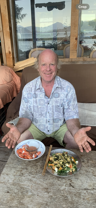 Un homme souriant est assis sur un canapé à l'extérieur et présente des assiettes de crevettes en sauce et une salade colorée. Il y a une table rustique en bois et un arrière-plan de lac.