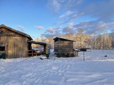 Eine verschneite ländliche Szene mit einer Holzscheune und einem kleineren Schuppen, umgeben von Zäunen und kahlen Bäumen. Helles Sonnenlicht und blauer Himmel schaffen eine friedliche Atmosphäre.