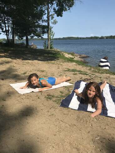 Zwei Mädchen in Badeanzügen liegen auf Handtüchern an einem Sandstrand am See. Bäume spenden Schatten, während eine andere Person in ein Handtuch gewickelt am Wasser sitzt.