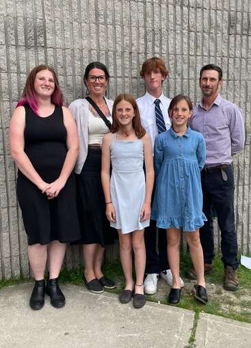 Six people, three adults and three children, stand smiling in front of a textured gray brick wall, dressed in casual and semi-formal outfits, posing together.