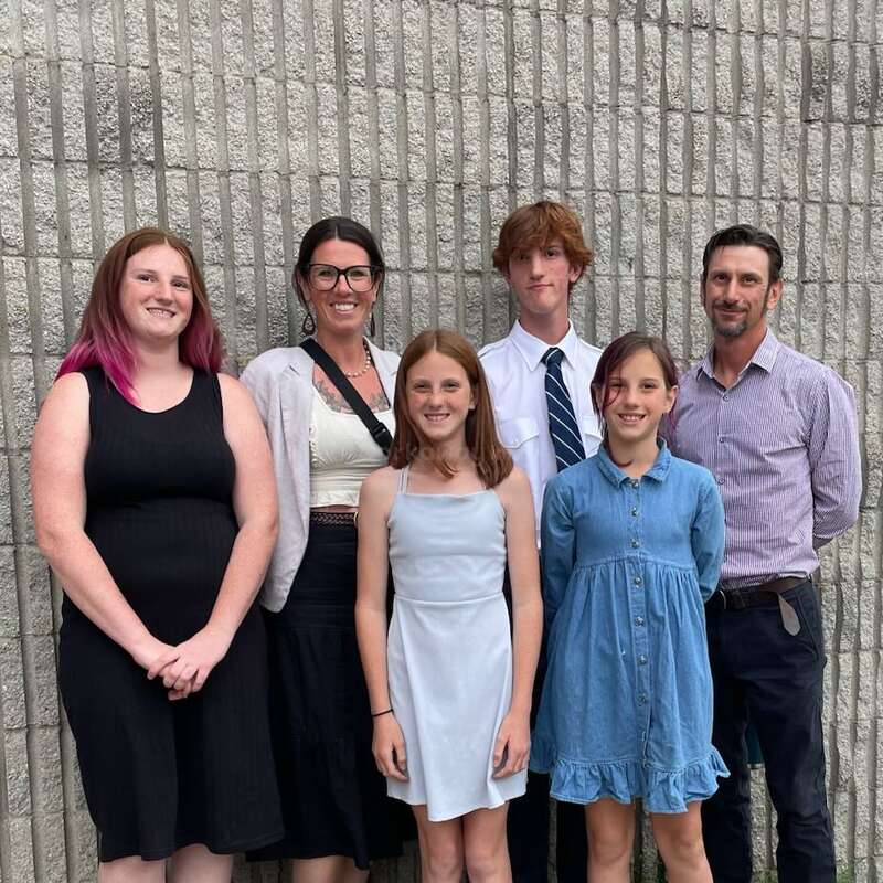 Sechs Personen, drei Erwachsene und drei Kinder, stehen lächelnd vor einer strukturierten grauen Backsteinmauer, gekleidet in legeren und halb-formellen Outfits, und posieren zusammen.