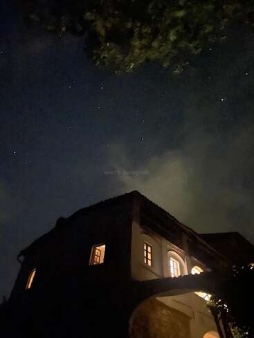 Une charmante maison aux fenêtres chaleureusement éclairées se trouve sous un ciel nocturne étoilé. De faibles nuages et une branche d'arbre ombragée ajoutent à l'atmosphère tranquille et mystérieuse.
