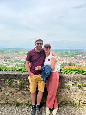 Un couple souriant se tient ensemble devant un point de vue panoramique, avec un paysage verdoyant et la ville en contrebas. Ils ont l'air heureux et détendus en plein air.
