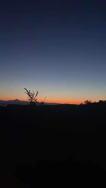 Une scène sereine de crépuscule avec un ciel dégradé passant du bleu profond à l'orange chaud, des montagnes silhouettées, des collines paisibles et un buisson solitaire au premier plan.