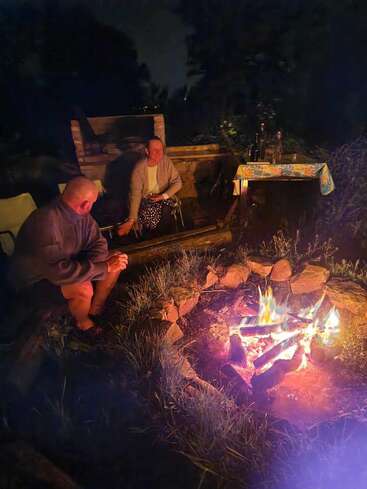 Deux personnes sont assises près d'un feu de camp rougeoyant la nuit, entourées de pierres, et discutent chaleureusement. Une table avec des bouteilles et une structure en briques se trouvent à l'arrière-plan.