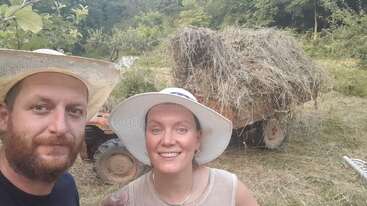Un homme et une femme portant des chapeaux sourient pour un selfie en plein air près d'un tracteur transportant un gros tas de foin, entouré de verdure et d'arbres.