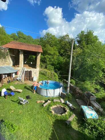 Un groupe de personnes profite d'une petite piscine hors sol dans une arrière-cour rustique, entourée de chaises, d'un vieux bâtiment en pierre, d'un foyer, d'une végétation luxuriante et du soleil.
