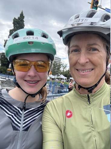 Dois ciclistas sorridentes tiram uma selfie ao ar livre, usando capacetes e camisetas de ciclismo. O dia está nublado e há árvores, tendas e pessoas ao fundo.