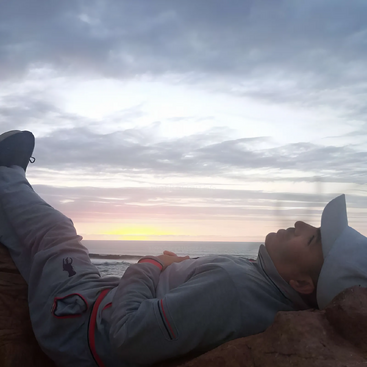 La imagen representa a una persona recostada en una roca, mirando al océano, con la cabeza apoyada en otra roca, con sombrero y ropa gris, en un cielo nublado.