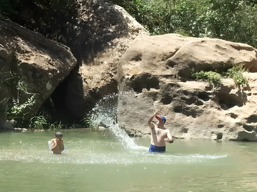 Zwei Jungen spielen und planschen in einem natürlichen Pool, umgeben von Felsen und Grünzeug. Das Sonnenlicht scheint und fängt einen lebhaften Sommermoment in der Umarmung der Natur ein.