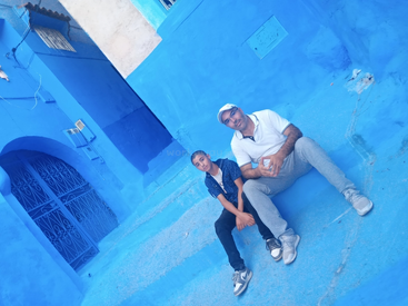 Ein Mann und ein Junge sitzen auf blau gestrichenen Stufen in einer lebhaften Straße mit blauen Wänden, wahrscheinlich in Chefchaouen, Marokko, lächeln und genießen die bunte Umgebung.