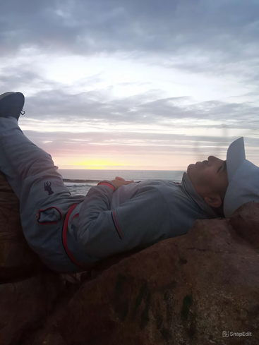 Das Bild zeigt eine Person, die auf einem Felsen liegt und auf das Meer blickt. Ihr Kopf ruht auf einem anderen Felsen, sie trägt einen Hut und graue Kleidung vor einem bewölkten Himmel.