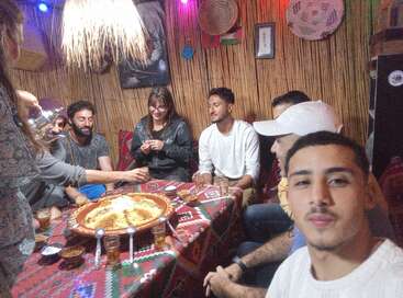 A group of friends sits around a table sharing a traditional meal, smiling and enjoying tea in a cozy, warmly decorated room with cultural decor.