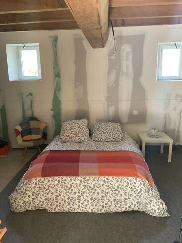 Um quarto aconchegante com uma cama de casal, roupa de cama floral e um cobertor laranja. Paredes sem acabamento, vigas de madeira no teto, duas janelas, cadeira de patchwork e uma mesa lateral.