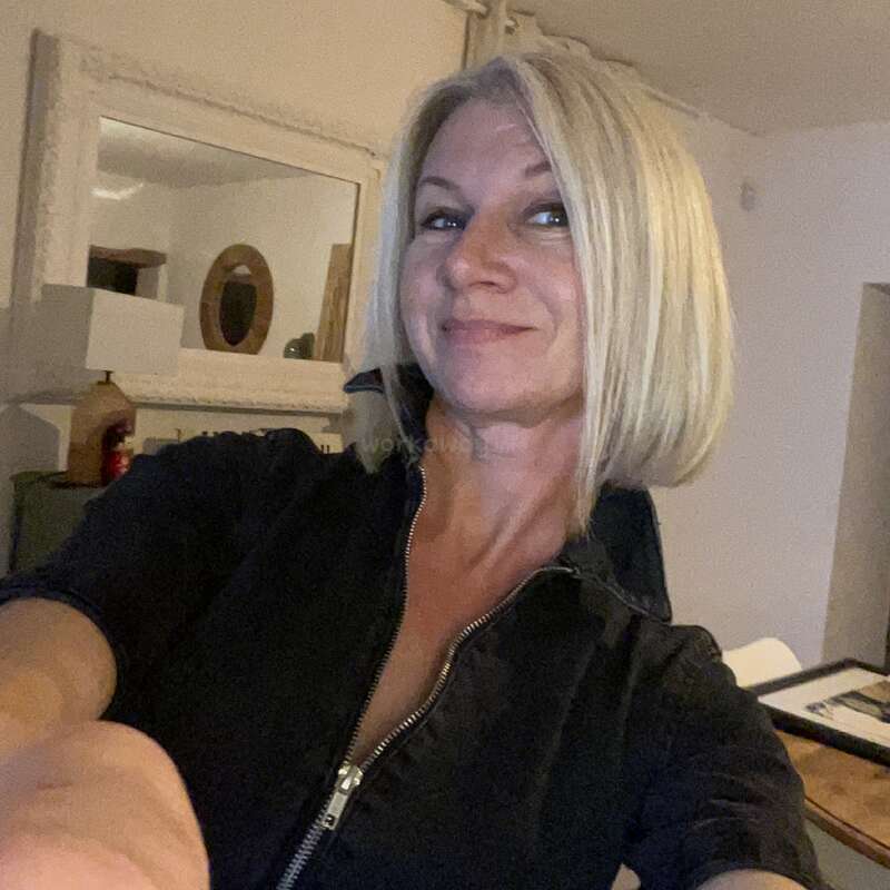 Uma mulher de cabelos loiros curtos está tirando uma selfie dentro de casa. Ela está sorrindo levemente, vestindo uma blusa escura com zíper, e há um espelho atrás dela.