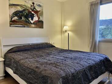 Cette chambre confortable comprend un grand lit avec une couverture grise texturée, un lampadaire moderne, un tableau de tauromachie coloré, des murs blancs, une fenêtre et une vue pittoresque sur la campagne.
