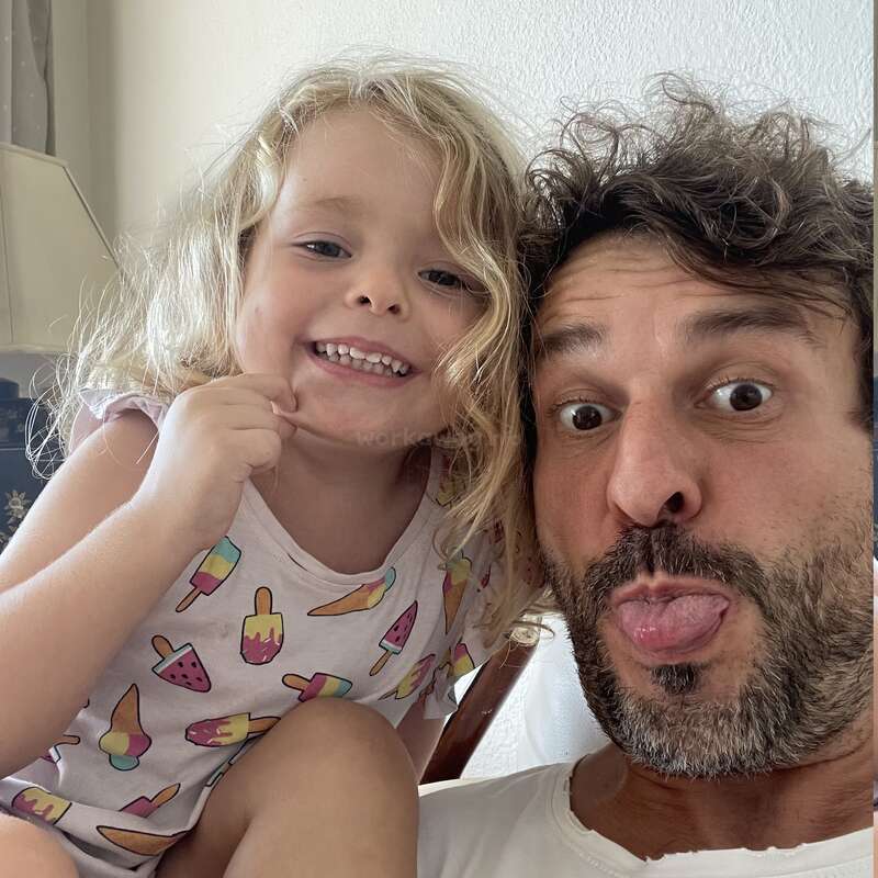 Une petite fille souriante aux cheveux blonds est assise à côté d'un homme barbu qui fait une drôle de grimace. Ils ont l'air enjoués et heureux, profitant ensemble d'un moment de légèreté.