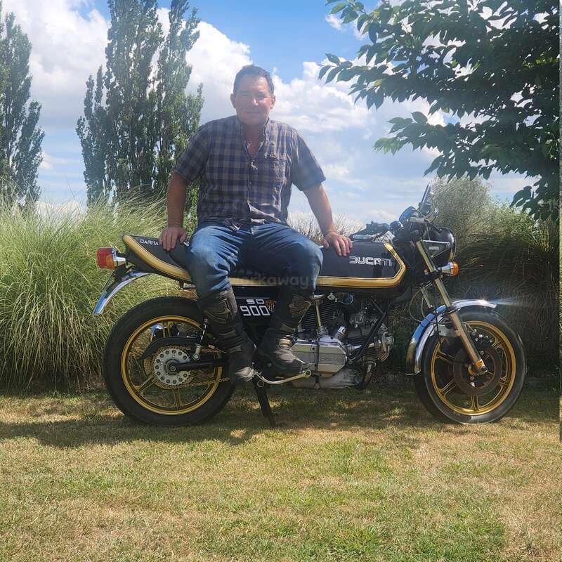A man in a plaid shirt and jeans sits on a vintage Ducati 900 motorcycle. He smiles, surrounded by tall grass, trees, and blue sky.