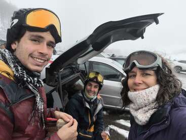 Trois personnes vêtues de tenues d'hiver et portant des lunettes de ski sourient près d'un coffre de voiture ouvert. Il neige et il y a du brouillard, ce qui laisse penser qu'elles font une pause après avoir skié ou fait du snowboard.