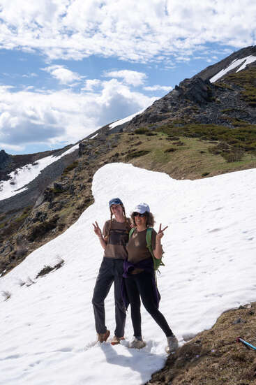 Deux femmes se tiennent sur un versant enneigé, souriantes, faisant le signe de la paix. Elles portent une tenue de randonnée sous un magnifique ciel bleu parsemé de nuages.