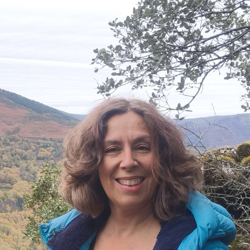 Une femme aux cheveux bruns bouclés sourit en plein air, vêtue d'une veste bleue. Derrière elle, on aperçoit des collines verdoyantes, des arbres et un ciel nuageux, formant un décor paisible et pittoresque.