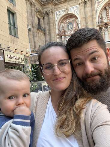 Une famille heureuse prend un selfie tous ensemble. La mère, le père et le bébé sourient devant un bâtiment historique richement décoré, orné de grandes colonnes et d'arcades.