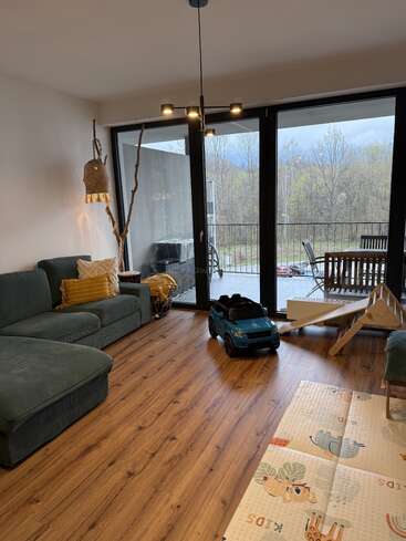 Ce salon confortable est équipé d'un canapé d'angle vert, de parquet, de jouets pour enfants, de grandes fenêtres donnant sur un balcon et offre une vue panoramique sur les arbres.