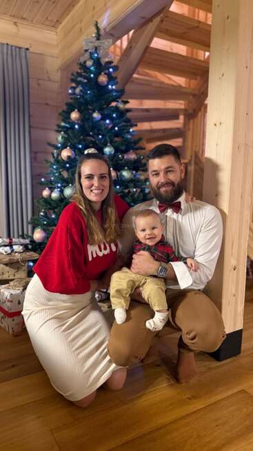 Uma família feliz posa em frente a uma árvore de Natal enfeitada dentro de uma aconchegante casa de madeira, sorrindo calorosamente, com presentes embrulhados no chão ao lado.