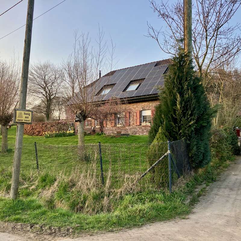 Uma casa de tijolos com painéis solares no telhado fica atrás de um quintal cercado, cercada por árvores e vegetação. Um carro branco está estacionado nas proximidades.