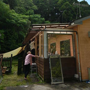 L'image représente une maison avec un extérieur en bois et un porche vitré, où deux personnes sont occupées à des travaux de construction ou de réparation sur le toit.