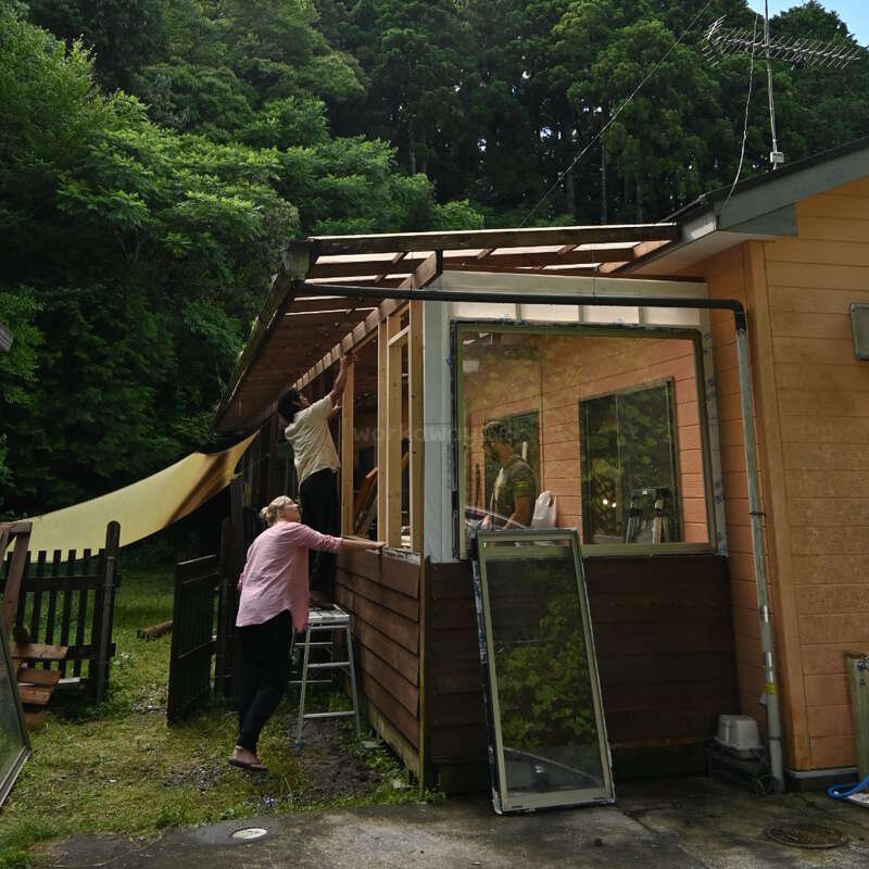 La imagen representa una casa con exterior de madera y porche acristalado, en la que dos individuos realizan trabajos de construcción o reparación en el tejado.