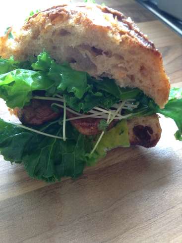 The image depicts a sandwich with a toasted bun, lettuce, sprouts, and meat, placed on a wooden surface, likely a table or cutting board.