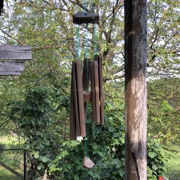 Un carillon en bambou est suspendu sous un porche et se balance doucement au-dessus des buissons et des arbres verdoyants, par une journée paisible et ensoleillée à la campagne.