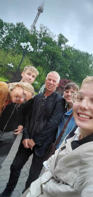 Un groupe de cinq personnes souriantes prend un selfie en plein air par un jour de pluie, avec des arbres verts et une haute tour visibles en arrière-plan.