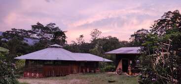 Una escena serena al atardecer muestra dos edificios rústicos de madera rodeados de frondosos árboles y vegetación. El cielo de tonos morados suaves y las flores crean una atmósfera apacible y tranquila.