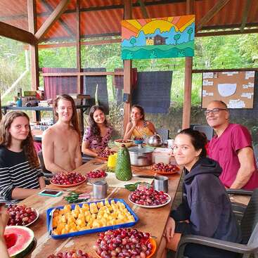 L'image représente un groupe de six personnes réunies autour d'une table chargée d'un assortiment de fruits, sur fond de paysage extérieur serein.
