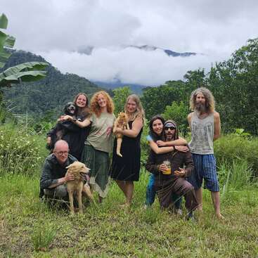 Un alegre grupo formado por seis adultos y tres mascotas posa al aire libre, rodeado de una exuberante vegetación y montañas bajo un cielo nublado, irradiando calidez y unión.