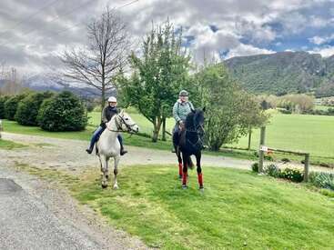 Duas pessoas andam a cavalo em uma margem de grama, usando capacetes, com uma estrada de cascalho, árvores e montanhas ao fundo, sob um céu nublado.