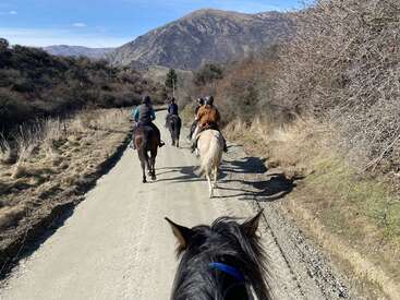 A imagem mostra um grupo de pessoas andando a cavalo em uma estrada de terra, cercado por árvores e montanhas, com um céu azul claro acima.