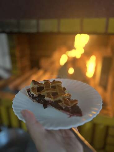 Uma mão segura um prato com uma fatia de torta de cereja em frente a uma lareira acesa, criando um ambiente aconchegante e acolhedor, perfeito para saborear uma sobremesa.