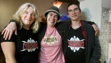 Three smiling people pose together indoors. Two wear black "Circus Palace" shirts, one wears a pink variant. The middle person sports a hat; all look happy and close.