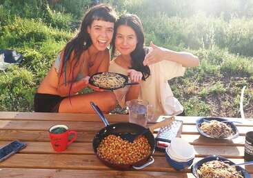 Two women sit at a wooden picnic table outdoors, smiling and presenting plates of pasta. Sunlight shines brightly. The table holds food, mugs, and a phone.
